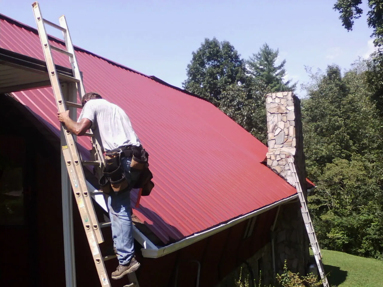 Roofer on a ladder beside a red metal roof and stone chimney — Soffit & Fascia Repair by Roofing Brothers in San Lorenzo
